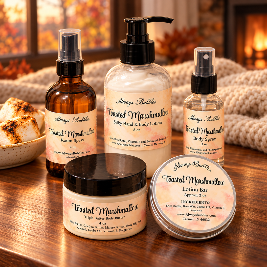 Always Bubbles Toasted Marshmallow gift set with lotion, 4 oz triple body butter, body spray, room spray, and lotion bar on a wooden table beside toasted marshmallows, with a knit blanket and fireplace in the autumn background.